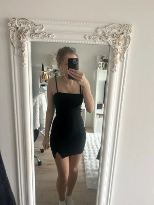 Svart kort klänning med slits - En svart, tight och kort klänning med smala axelband och slits på sidan. Klänningen har en enkel och stilren design som framhäver figuren. Perfekt klänning för fest. 