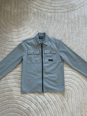 Grå overshirt från Closure - Helt ny använd 2 gånger endast. Stilren grå overshirt från Closure med rak passform och svarta detaljer. Jackan har två bröstfickor med knappar, klassisk krage och en svart dragkedja framtill. Materialet är slitstarkt och passar perfekt för lager-på-lager. Diskret logopatch på bröstet. Köptes in för 1200kr 