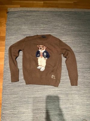Brun stickad tröja med björn från Ralph Lauren - Säljer en brun stickad tröja från Ralph Lauren med ikonisk björn på bröstet. Björnen har blå collegejacka, randig tröja och vita byxor. RL-broderi nertill. Rund hals och ribbade muddar. Perfekt för dig som gillar preppy stil.