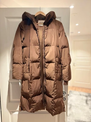Brun lång dunjacka från Mos Mosh - Snygg brun lång dunjacka från Miss Miss med huva och quiltad design. Jackan har dragkedja framtill, två sidofickor och en patch med logga på ärmen. Perfekt för kalla vinterdagar och ger en riktigt trendig look.