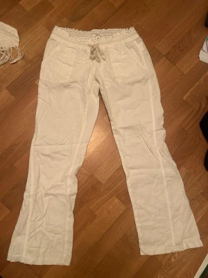 Vita linnebyxor från Roxy - Säljer ett par vita linnebyxor från Roxy med avslappnad passform, resårmidja och dragsko i midjan. Byxorna har två framfickor och är tillverkade i ett lätt och luftigt linnematerial, perfekta för en chill och stilren look. Bootcut, lågmidjade