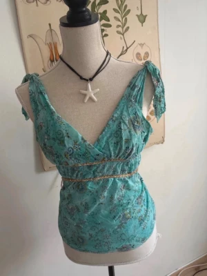 Turkos blommig omlottopp med knyt - Snygg turkos omlottopp med blommigt mönster och knyt på axlarna. Toppen har v-ringning och markerad midja med dekorativt band. Perfekt för dig som gillar bohemisk stil och vill ha något lätt till sommaren.