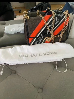 Michael Kors handväska med kedja - Snygg handväska från Michael Kors i brunt konstläder med orange och svarta detaljer samt stora loggor. Väskan har guldfärgade kedjehandtag och ett vridlås framtill. Perfekt accessoar för dig som vill sticka ut med en trendig och färgstark look.