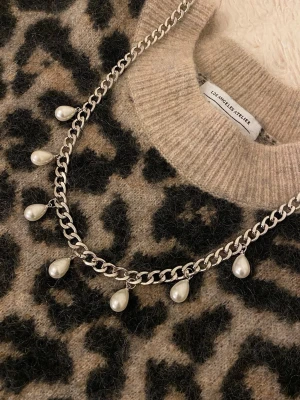 Silverfärgat halsband med pärldroppar - Stilrent halsband i silverfärgad metall med sju hängande droppformade pärlor. Kedjan är grov och ger ett trendigt intryck, perfekt för att lyfta din outfit med en cool detalj. Pärlorna är vita och ger en snygg kontrast mot den silvriga kedjan.