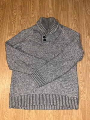 Grå stickad tröja med hög krage och knappar - Snygg och bekväm grå stickad tröja med hög ribbad krage och dekorativa knappar framtill. Mjuk och varm – perfekt för höst och vinter. Ribbstickade muddar vid ärmslut och nederkant ger en fin passform.  Passar lika bra till jeans för en avslappnad look som till chinos för en mer uppklädd stil.  Bra skick. Hör gärna av dig vid frågor eller för fler bilder 😊