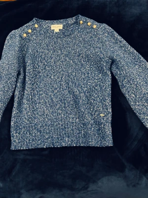 Holly and white stickad tröja - Blåmelerad stickad tröja sopuffade axlar från holly&white. Tröjan har fyra guldfärgade knappar på varje axel, rund halsringning och ribbade muddar. Materialet är en mjuk blandning av bomull och ull. Perfekt för dig som gillar klassisk men trendig stil.