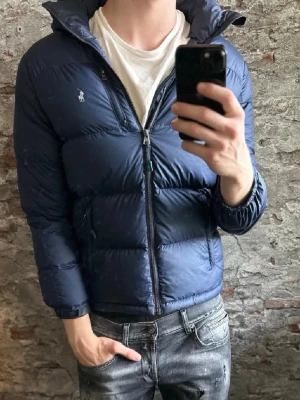 Mörkblå dunväst från Polo Ralph Lauren - Snygg mörkblå dunväst från Polo Ralph Lauren med hög krage och dragkedja framtill. Västen har två sidofickor med dragkedja och den klassiska loggan broderad på bröstet. Perfekt för lager på lager och ger en sportig vibe.