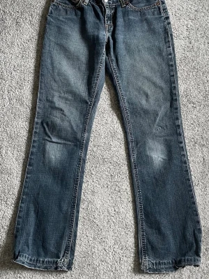 Levi's blå bootcut jeans - Säljer ett par klassiska blå Levi's jeans med bootcut passform. Jeansen har slitna detaljer vid knän och benslut, fem fickor och synliga sömmar. Tillverkade i denim med en autentisk tvättad look och läderpatch bak i midjan.