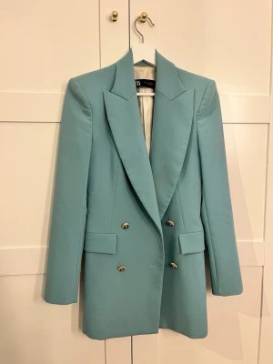 Ljusblå kostym från Zara - Säljer en stilren ljusblå kostym från Zara med dubbelknäppt kavaj och matchande byxor med pressveck. Kavajen har breda slag, guldknappar och fickor med lock. Byxorna är raka och har hög midja. Perfekt för dig som vill sticka ut med färg! Kavaj storlek Xs och byxor storlek S