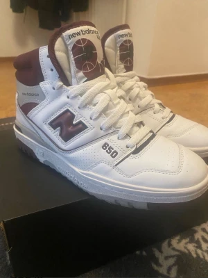 New Balance 650 vita/bordeaux sneakers - Snygga New Balance 650 sneakers i vitt och bordeaux med högt skaft. Skorna har klassisk basketinspirerad design, perforerad tå och tydliga loggor på sidan och plösen. Tillverkade i läder med detaljer i textil och gummi. Perfekta för dig som gillar retrostil.