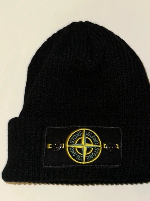 Svart ribbstickad mössa Stone Island - Svart ribbstickad mössa från Stone Island med den klassiska logotypen framtill i gult och grönt på en svart patch. Mössan har uppvikt kant och är tillverkad i ett mjukt, värmande material. Perfekt för dig som vill ha en stilren och ikonisk accessoar.