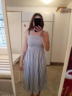 Randig blå och vit långklänning - Säljer en somrig långklänning med smock upptill och smala axelband. Klänningen har ett randigt mönster i blått och vitt, och är tillverkad i ett lätt bomullstyg som är perfekt för varma dagar. Passformen är normal och klänningen har en luftig och avslappnad stil.