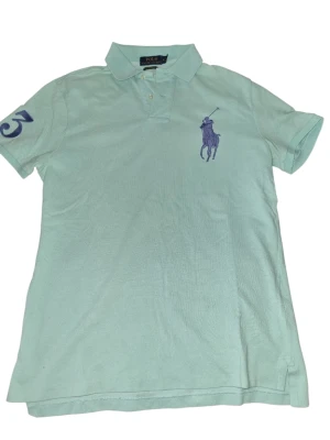 Mintgrön pikétröja från Ralph Lauren - Snygg mintgrön pikétröja från Ralph Lauren med klassisk krage, knappar framtill och stor lila logga på bröstet. Tröjan har korta ärmar och en broderad siffra på ena ärmen. Perfekt för en avslappnad och stilren look.