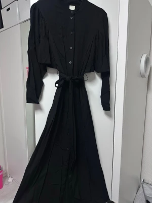 Svart långklänning med knappar och bälte - Stilren svart långklänning med knappar framtill och matchande bälte i midjan. Klänningen har långa ärmar och en rak, elegant siluett. Perfekt för dig som gillar minimalistisk och tidlös stil.