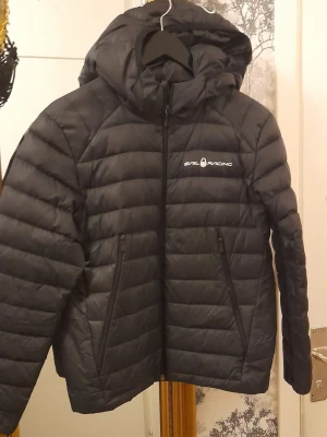 Sail Racing Jr Spray Down Dunjacka  - Snygg svart dunjacka från Sail Racing med huva och dragkedja framtill. Jackan har två praktiska sidofickor med dragkedja och diskret logga på bröstet. Quiltad design som ger en modern look och håller dig varm under kalla dagar. Stl 150