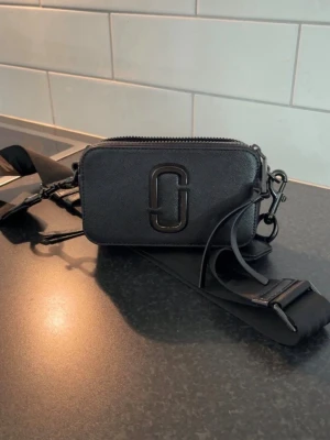 Svart Marc Jacobs Snapshot handväska - Säljer en svart Marc Jacobs Snapshot handväska i rektangulär form med bred axelrem och dubbla dragkedjor. Väskan har en stor logga framtill och är gjord i tåligt läder. Perfekt för dig som gillar stilrena accessoarer med en modern touch.