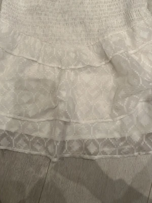 Vit kortkjol med volang och mönster - Supersöt vit kortkjol med volangdetaljer och diskret geometriskt mönster. Kjolen har smock i midjan för en bekväm passform och är gjord i ett lätt tyg som passar perfekt till varmare dagar.