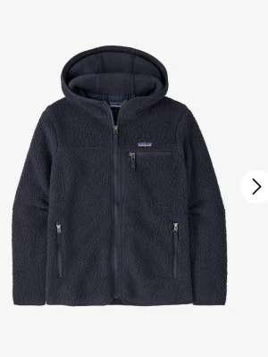 Mörkblå teddyjacka från Patagonia - Mysig mörkblå teddyjacka från Patagonia med huva och hel dragkedja. Jackan har tre fickor med dragkedja, varav en på bröstet, och är tillverkad i fleece-material. Perfekt för kalla dagar och har en normal passform.