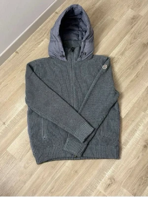 Moncler cardigan - Snygg grå cardiganjacka från Moncler med stickade ärmar och framstycke samt quiltad huva och rygg. Dragkedja framtill och två fickor. Moncler-logga på ärmen. Perfekt för dig som vill ha både stil och komfort.