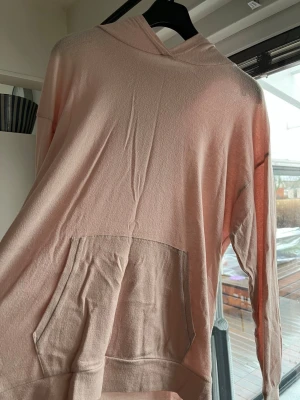 Ljusrosa hoodie med magficka - Mysig ljusrosa hoodie med stor magficka och huva. Hoodien har en avslappnad passform och är tillverkad i ett mjukt tyg, perfekt för en chill look. Långa ärmar och ribbad nederkant ger en klassisk hoodie-stil.