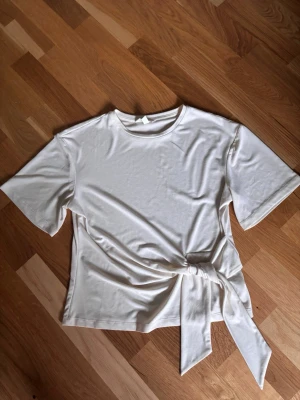 Vit topp med knyt i midjan - Stilren vit topp med rund halsringning och korta ärmar. Toppen har en snygg knyt-detalj i midjan som ger en trendig touch. Perfekt att matcha med jeans eller kjol för en enkel men chic look.