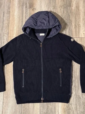 Moncler cardigan  - Snygg svart hybridjacka från Moncler med stickade ärmar och quiltad kropp i polyester. Jackan har huva, dragkedja framtill och två fickor med dragkedja. Perfekt för dig som vill ha en modern och sportig look.