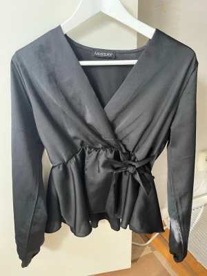 Svart omlottblus från Missay - Stilren svart omlottblus från Missay med långa ärmar och knytband i midjan. Blusen har en lätt glansig finish och volang nedtill som ger en feminin touch. Perfekt för att lyfta vilken outfit som helst.