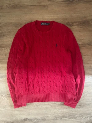 Röd kabelstickad tröja Polo Ralph Lauren - Kabelstickad röd tröja från Polo Ralph Lauren med rund halsringning och klassisk marinblå logga broderad på bröstet. Tröjan har ribbade muddar vid ärmslut och nederkant. Perfekt för dig som gillar tidlös och preppy stil. 100% äkta och storlek M. Tröjan har en liten fläck längst ner som förmodligen går bort med en tvätt.