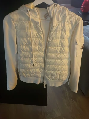 Monclear jacka använd 1 gång - Snygg vit dunjacka från Moncler med quiltad front, huva med dragsko och ribbade muddar. Jackan har dragkedja framtill och en ficka med lock och Moncler-logga på ärmen. Perfekt för kalla vinterdagar och riktigt trendig stil.
