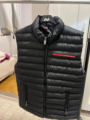 Svart dunväst från Prada - Säljer en svart Prada väst som är helt ny och oanvänd. Stilren och tidlös modell med dragkedja framtill samt två sidofickor med dragkedja. Röd Prada-logo framtill och exklusiv Prada Milano på insidan.