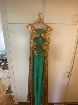 Grön och guldglittrig långklänning - Snygg långklänning i grönt tyg med breda guldglittriga paneler längs sidorna. Klänningen har unika utskärningar framtill och är ärmlös. Perfekt för dig som vill sticka ut med en glamorös look.