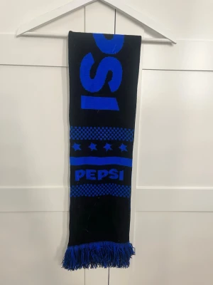 Svart och blå halsduk med Pepsi-logo - Snygg svart halsduk med blått Pepsi-tryck och stjärnmönster. Halsduken har fransar i ändarna och är tillverkad i ett mjukt, stickat material. Perfekt för dig som gillar retro eller vill sticka ut med en unik accessoar.