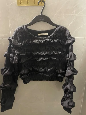 Svart croppad topp från NA-KD - Säljer en svart croppad topp från NA-KD med långa ärmar och volangdetaljer. Toppen har en glansig finish och är tillverkad i ett syntetmaterial som ger en cool look.