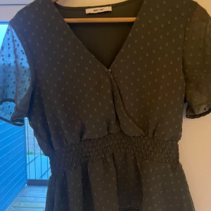 Grön blus med prickigt mönster från About You - Supersöt grön blus från About You med små prickar och markerad midja med smock. Blusen har korta ärmar i transparent tyg och v-ringad hals. Perfekt för vår och sommar!