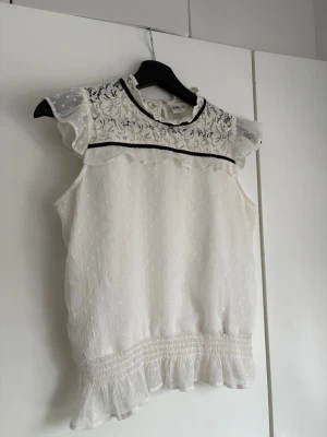 Vit spetsblus med volangdetaljer - Supersöt vit blus med spets upptill och volangdetaljer vid ärm och nederkant. Blusen har svarta banddetaljer och är ärmlös. 