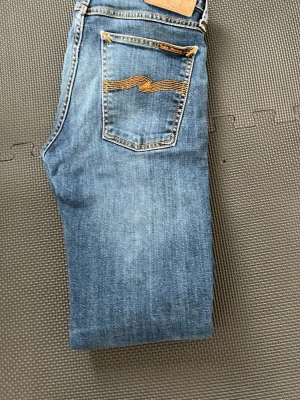 Blå jeans från Nudie Jeans - Snygga blå jeans från Nudie Jeans med klassisk femficksdesign och gul brodyr på bakfickan. Jeansen har raka ben och är tillverkade i denim med tydliga slitningar för en cool look.