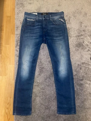 Blå slim fit jeans från Replay - Snygga blå slim fit jeans från Replay med klassisk femficksdesign och diskreta slitningar framtill. Jeansen har en mörk tvätt med ljusare partier på lår och knän samt svarta detaljer vid fickorna. Perfekta för en trendig och avslappnad look.