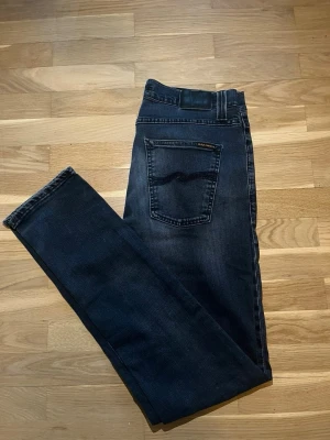 Grå/svarta jeans från Nudie Jeans - Snygga mörkgråa jeans från Nudie Jeans med klassisk femficksmodell och subtila slitningar framtill. Jeansen har raka ben och en diskret logga på bakfickan. Tillverkade i mjuk denim som ger en skön passform och stilren look. Storlek 32/34
