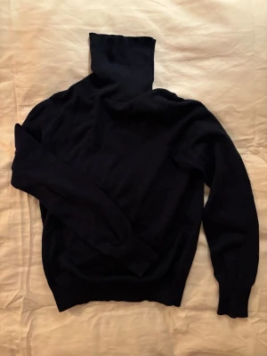 Mörkblå polotröja i mjukt stickat material - Säljer en mörkblå stickad polotröja med lång ärm och ribbade muddar. Tröjan har en hög polokrage och är tillverkad i ett mjukt, tunt material som känns skönt mot huden. Perfekt för lager på lager och enkel att matcha med olika outfits.