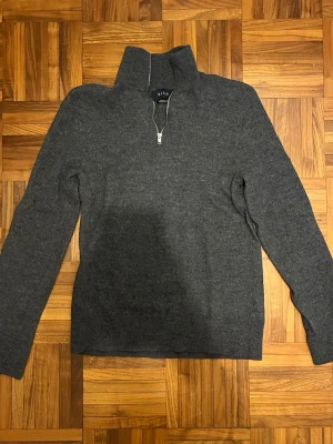 Mörkgrå stickad halvzip från Riley - Mörkgrå stickad tröja från Riley med halvzip och hög krage. Tröjan har långa ärmar och är tillverkad i ett mjukt material som passar perfekt för kyligare dagar. Enkel och stilren design som funkar till många outfits. Krympt från S till xs mått i bilderna