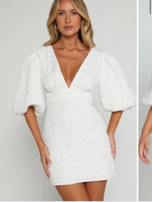 Vit kort klänning med puffärmar - Snygg vit kort klänning med v-ringning och puffiga kortärmar. Klänningen har en markerad midja och är tillverkad i ett strukturerat tyg med subtilt mönster. Perfekt för dig som vill ha en trendig och elegant look.