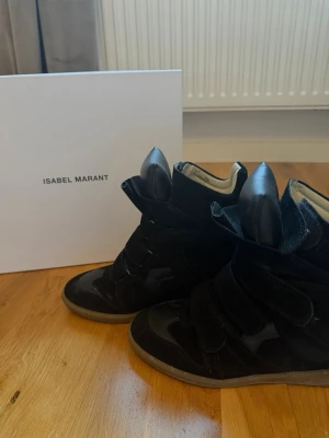 Isabel Marant svarta höga sneakers - Säljer ett par svarta höga sneakers från Isabel Marant i mocka med coola detaljer och kardborreband. Dem är använda max 10 ggr inget av mockan är skadat utan som ni ser på bild 2 bara det bruna är slitet pga man använt dem ute då men inget som man tänker på eller syns när man har dem. Dem är köpte nya på NK för 5900kr.