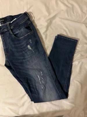 Blå slitna jeans från Replay - Säljer ett par blå jeans från Replay med snygga slitningar och tvättade detaljer. Jeansen har klassisk femficksmodell, normal midja och smal passform. Perfekta för dig som gillar en avslappnad men trendig stil.