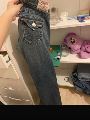 B jeans från True Religion - Säljer ett par blå jeans från True Religion med klassiska kontrastsömmar och stor ficka bak med lock och knapp. Jeansen har rak men ser bootcut på  passform och är tillverkade i denim med detaljer i vitt och orange. Perfekta för dig som gillar streetwear och vill ha en ikonisk look.