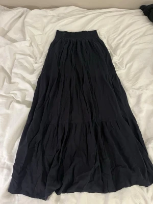 Svart långkjol med resårmidja - En svart långkjol med bred resår i midjan och mjukt, lätt tyg som faller snyggt. Kjolen har en enkel och flowy design utan mönster eller detaljer, perfekt för en avslappnad och stilren look.