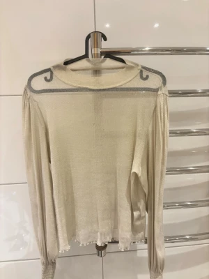 Glittrig ljus långärmad topp - Snygg ljusglittrig långärmad topp med transparenta detaljer vid axlarna och smockade muddar. Materialet är tunt och skimrande, vilket ger en elegant look. Perfekt för dig som vill sticka ut lite extra.