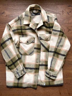 Jacka  - Stilren overshirt från Zara i mjuk ullmix med rutigt mönster i beige, olivgrönt, svart och ljusrosa. Två bröstfickor med lock och tryckknappar, klassisk krage och raka ärmar. Perfekt lager på lager-plagg för en trendig look.