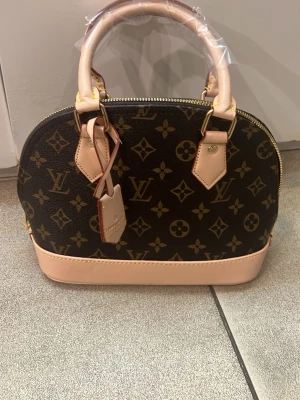  monogram handväska - Snygg handväska från Sanni  i klassiskt brunt monogrammönster med beigea detaljer och handtag. Väskan har guldfärgade metalldetaljer och dragkedja upptill. Perfekt för dig som vill ha en ikonisk och lyxig accessoar till din outfit.