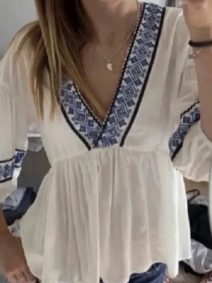 Vit blus med blå brodyr från Zara - Säljer en luftig vit blus från Zara med blått broderi längs V-ringningen och ärmarna. Blusen har en avslappnad passform, volang nedtill och långa ärmar med resår vid ärmslut. Tröjan finns ej kvar så därav pris💕