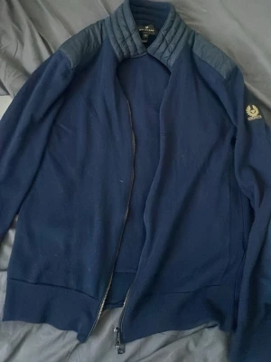 Mörkblå Belstaff cardigan - Mörkblå cardigan från Belstaff med quiltade detaljer på axlar och krage. Den  har dragkedja framtill och en broderad logga på ärmen. Tillverkad i mjukt material med ribbade muddar vid ärmslut och nederkant.